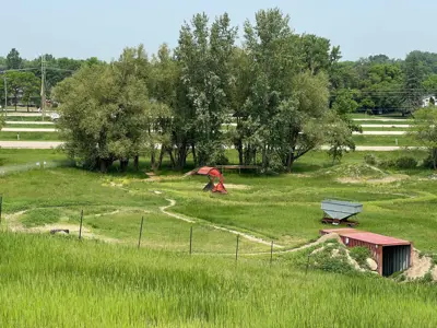 Grassy area at Prairie Flow Trails bike and activity park in Portage la Prairie, featuring a winding dirt path, red sculpture, and stacked metal containers near trees and a roadside